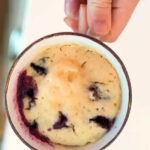 A hand holding a mug filled with a baked blueberry muffin in a mug cake, revealing juicy blueberries inside.