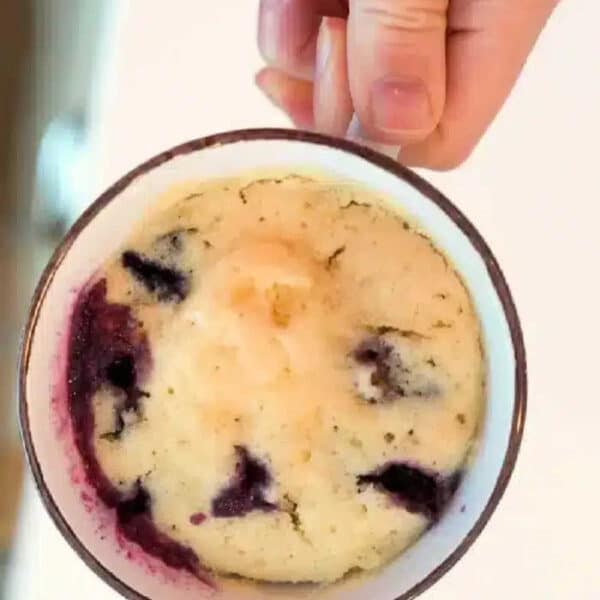 A hand holding a mug filled with a baked blueberry muffin in a mug cake, revealing juicy blueberries inside.