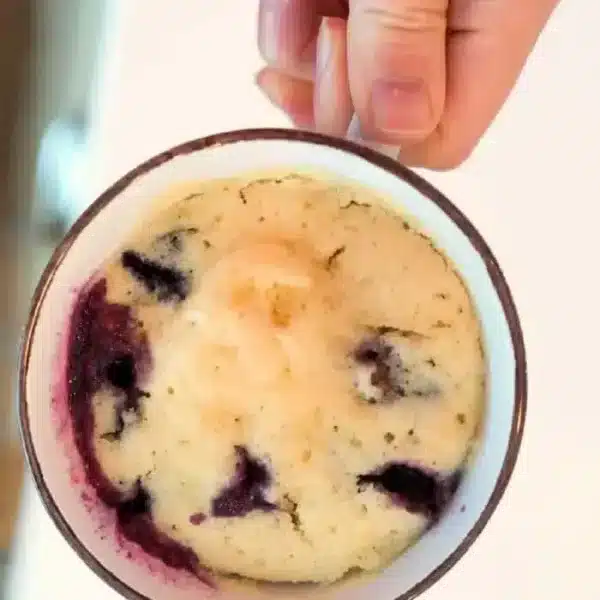 A hand holds a mug with a delightful blueberry muffin in a mug inside. The cake is light in color, dotted with juicy blueberries that have gently stained the sides. The mug sits elegantly on a pristine white surface.