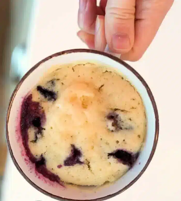 A hand holds a mug with a delightful blueberry muffin in a mug inside. The cake is light in color, dotted with juicy blueberries that have gently stained the sides. The mug sits elegantly on a pristine white surface.