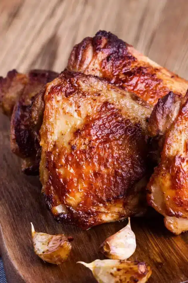 A close-up view of several pieces of golden-brown, bone-in air fryer chicken thighs with crispy skin on a wooden surface, accompanied by a few roasted garlic cloves. The deliciously well-cooked chicken promises a savory experience.