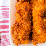 Close-up of crispy Cheese It Chicken Tenders, their orange coating glistening on a white plate, set atop a red and white striped towel.