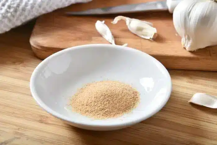 A small white dish containing garlic powder rests on a wooden surface, perfect for seasoning the best baked pork chops. Behind it, garlic cloves and a cutting board with a knife add to the culinary tableau.