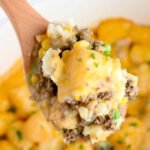 A wooden spoon holds a serving of crock pot tater tot casserole with cheesy ground beef, peas, and corn.