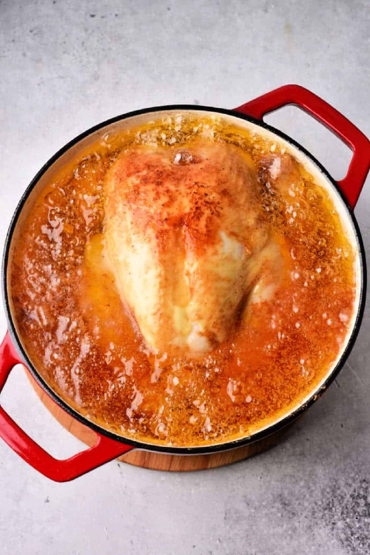 Whole chicken simmering in bubbling broth inside a red Dutch oven on a grey surface, ready to become the juiciest Whole Fried Chicken.