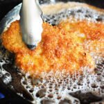 Tongs holding a golden-brown fried chicken cutlet sizzling in hot oil.