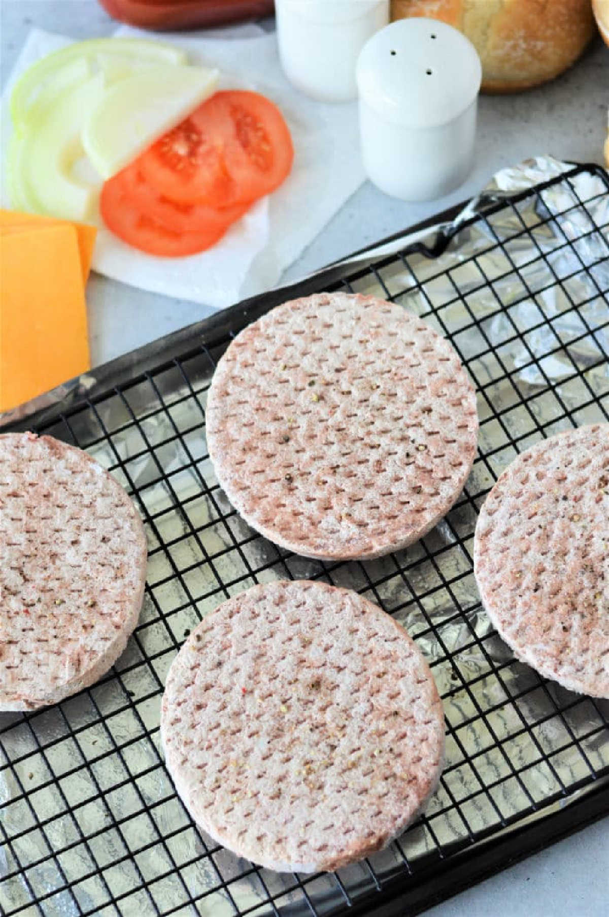 Four frozen hamburger patties on a wire rack, with sliced tomato, onion, cheese, and seasonings nearby&mdash;perfect for learning how to cook frozen burgers in the oven for a quick and tasty meal.