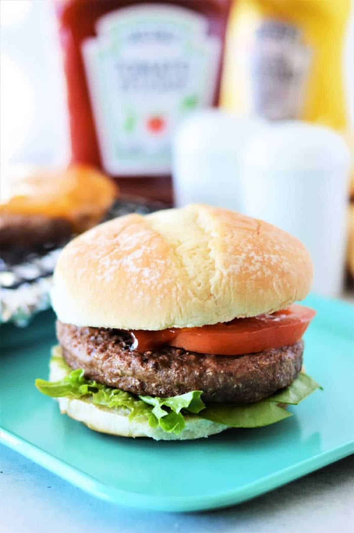 A hamburger with lettuce and tomato on a blue plate, with ketchup and mustard bottles in the background&mdash;perfect inspiration for learning how to cook frozen burgers in the oven for a quick, delicious meal.