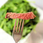A fork holds a slice of rare ribeye in air fryer steak above a blurred background of green leafy vegetables.
