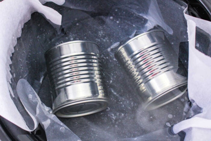 Two metal cans in boiling water inside a pot lined with parchment paper, demonstrating how to make caramel with condensed milk.