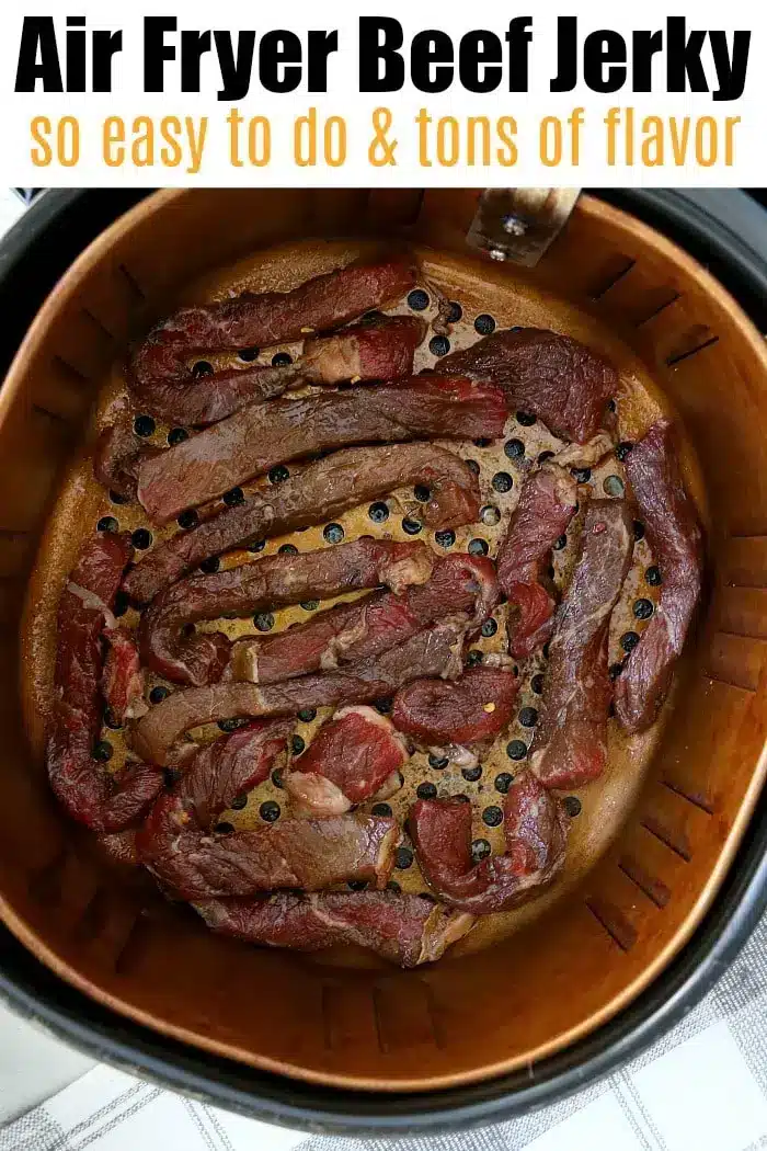 A metal air fryer basket is filled with pieces of savory beef jerky. The top of the image contains text that reads: "Air Fryer Beef Jerky so easy to do & tons of flavor." The setup is on a gray-patterned countertop.