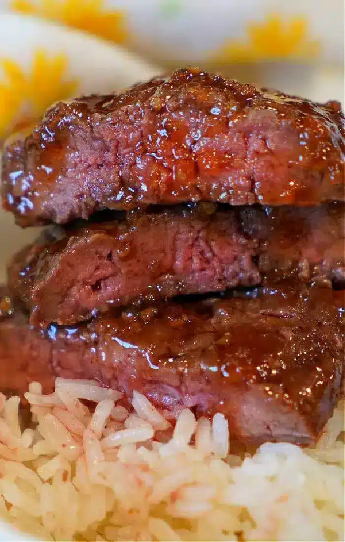 A close-up shows three slices of barbecued beef with a shiny glaze stacked on top of each other, placed on a bed of white rice. The meat appears tender and moist with a slightly charred exterior, reminiscent of popular air fryer beef recipes. The background hints at a colorful, patterned tablecloth.