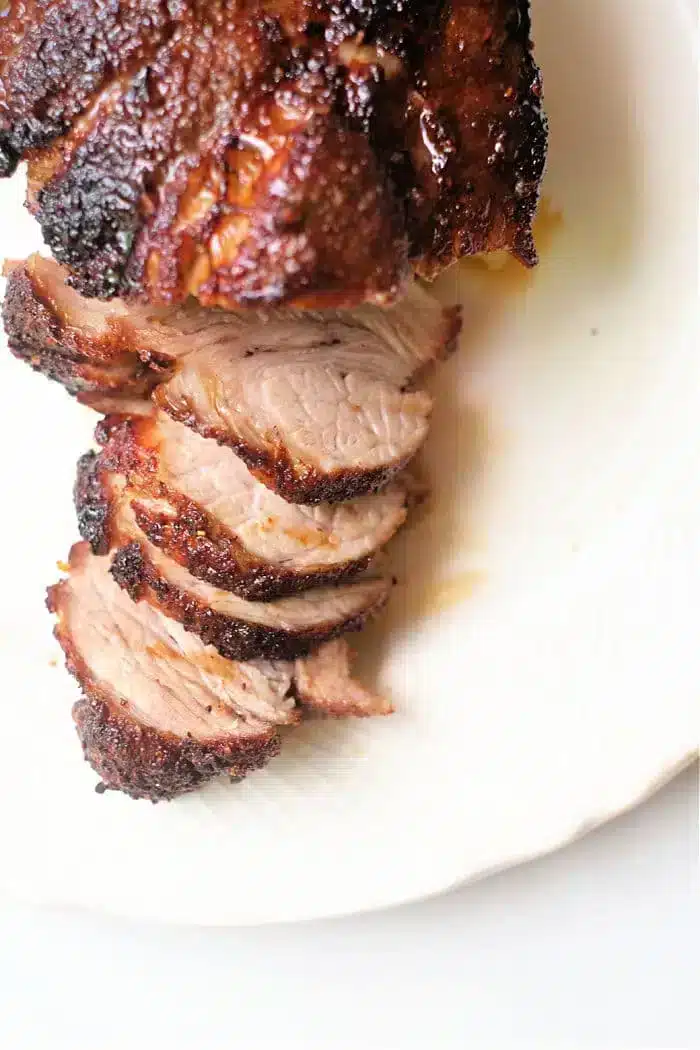 A close-up view of a roasted, sliced air fryer pork roast on a white plate. The meat is browned and crispy on the outside, with a juicy, tender interior. Five slices are visible, arranged neatly in a row.