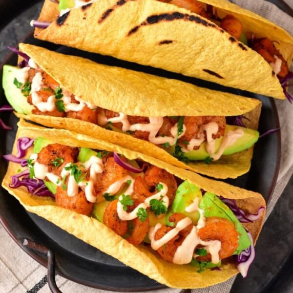 Three grilled corn tacos filled with shrimp, avocado, and cabbage, drizzled with creamy sauce on a dark plate—try making the taco shells in an air fryer for extra crispiness.