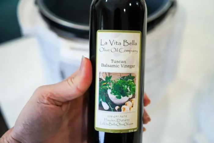 A hand holds a bottle of La Vita Bella Olive Oil Company's Tuscan Balsamic Vinegar, perfect for enhancing your beef arm roast recipe. The label features images of herbs and garlic, beautifully showcased against a blurred background.