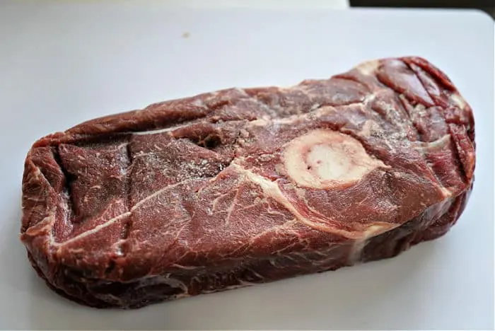 A raw cut of beef arm roast on a white surface, featuring a bone in the center. The meat has a marbled texture and a reddish-brown color, perfect for your next savory recipe.