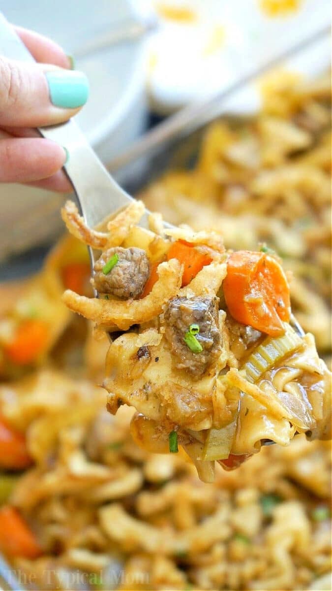 Easy beef stew casserole is comfort food at it's finest!! Packed with tender meat, a thick sauce and lots of vegetables you and your kids are sure to have seconds! Use stew meat or ground beef to make this simple casserole that may become your new family favorite meal. #beef #stew #casserole #easy #vegetable #groundbeef A fork holding a bite of easy beef stew casserole with noodles, beef, carrots, celery, and crispy onions.