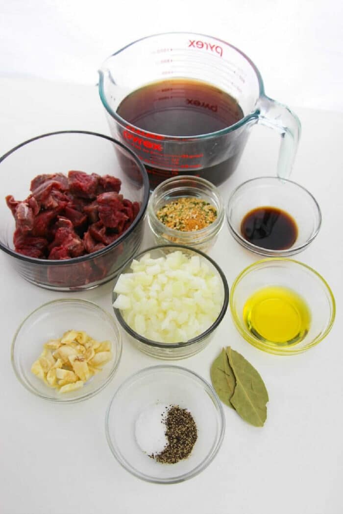 A selection of ingredients for how to cook beef tips, featuring cubed beef, broth, chopped onions, garlic, oil, spices, herbs, and soy sauce.