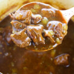 A wooden spoon lifts chunks of beef stew with vegetables from a pot of thick, rich broth—perfect inspiration for anyone learning how to cook beef tips.