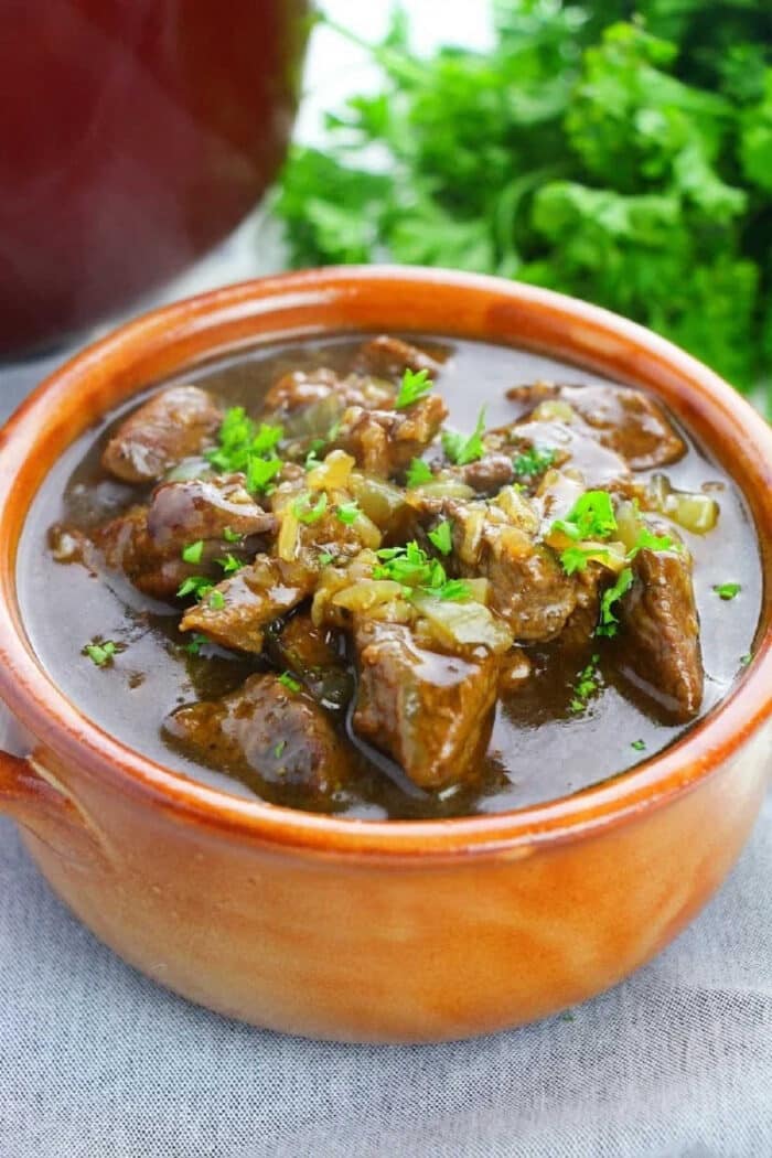 Brown ceramic bowl of beef stew garnished with chopped parsley, placed on a light cloth, with herbs in the background—a delicious result when you learn how to cook beef tips perfectly.