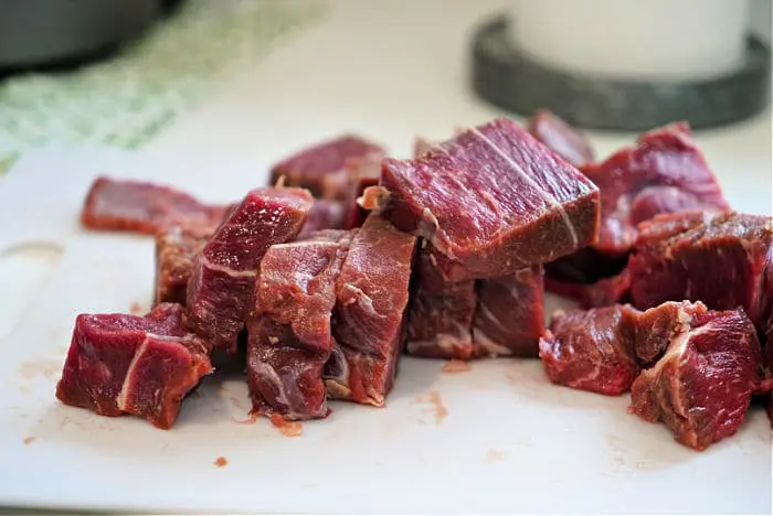 Chopped pieces of raw beef are placed on a white cutting board, perfect for starting a beef arm roast recipe. The meat is marbled with fat, and the board rests on a kitchen counter. The background features a green-patterned cloth and part of a round, gray object.