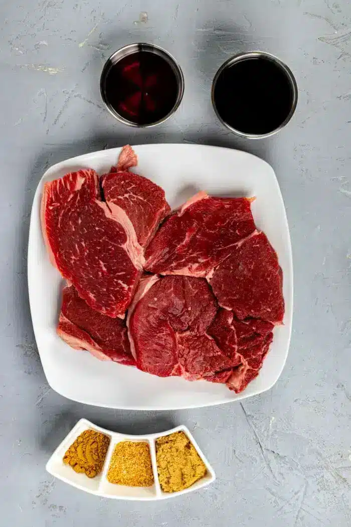 A square white plate with several pieces of raw red beef sits on a textured gray surface, suggesting a perfect base for an Asian steak marinade. Above the plate are two cups, one filled with a dark liquid and the other with a reddish liquid. Below the plate are three small white dishes with various spices.