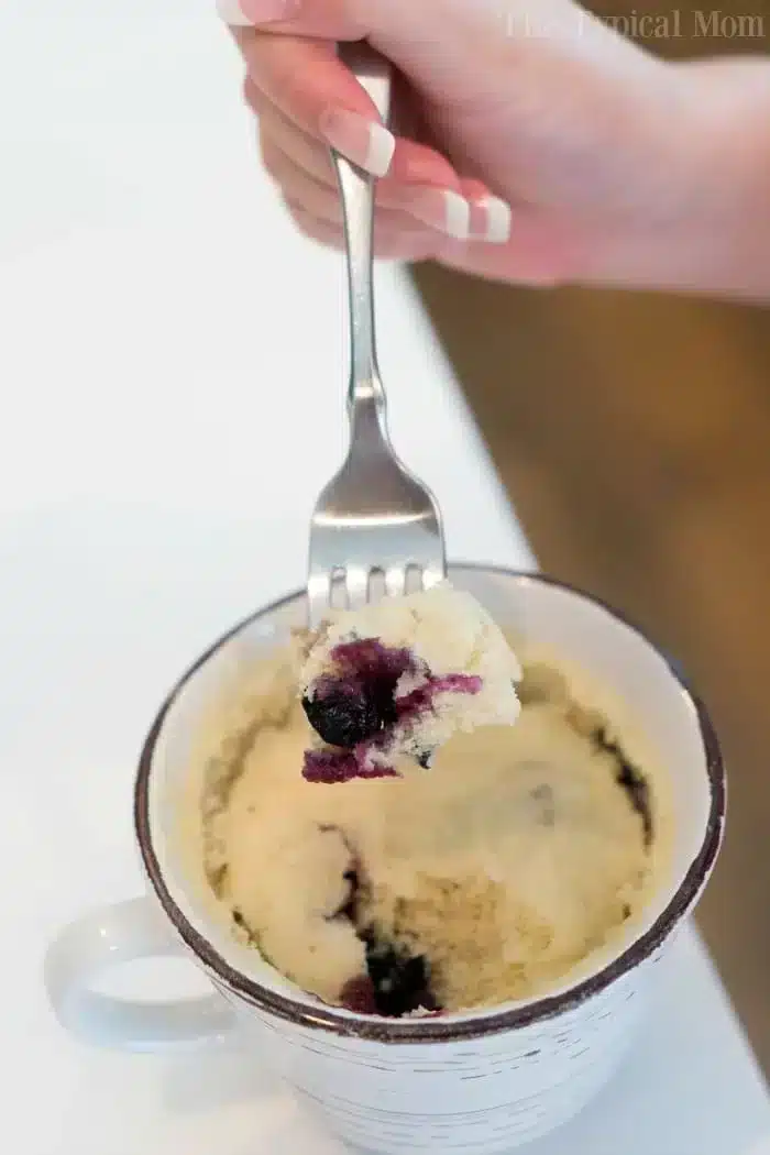 A hand holding a fork with a piece of blueberry muffin in a mug above a white ceramic mug filled with the cake. The mug is placed on a white surface.