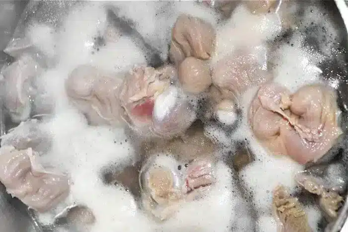 Chunks of raw chicken boiling in water, producing foam and bubbles on the surface. The chicken pieces appear light-colored and are partially submerged in the boiling liquid, reminiscent of preparing Air Fryer Chicken Gizzards with a traditional twist.