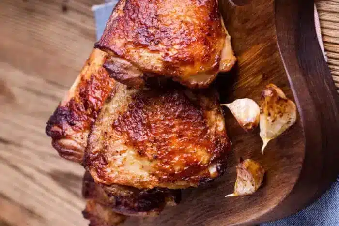 Grilled bone-in chicken thighs with crispy, browned skin are arranged on a wooden cutting board. Garlic cloves, also roasted, are placed next to the chicken pieces. The background features a wooden surface with a rustic appearance.