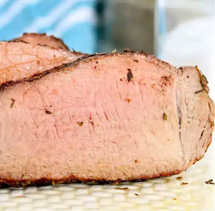 A close-up view of a thick, sliced piece of cooked meat with a pink interior and slightly charred outer edge, perfect for air fryer beef recipes. The background is slightly blurred, showing a blue and white striped cloth and a surface.