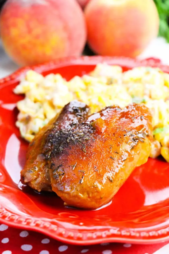 A plate with boneless skinless chicken thighs, glazed to perfection, alongside a side of macaroni salad on a red dish. Two peaches are blurred in the background.