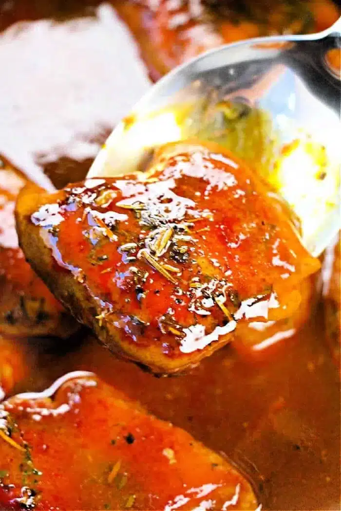 A close-up of a spoon lifting a piece of boneless, skinless chicken thigh coated in a glossy, reddish-brown sauce. The sauce appears thick and well-seasoned with visible herbs. More tender pieces simmer in the crockpot’s sauce in the background.