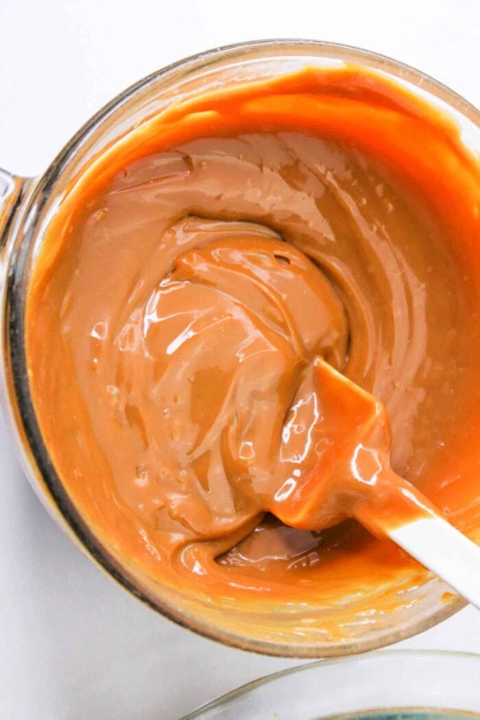 A bowl of melted caramel made from condensed milk is being stirred with a white spatula, showing how to make caramel with condensed milk at home.