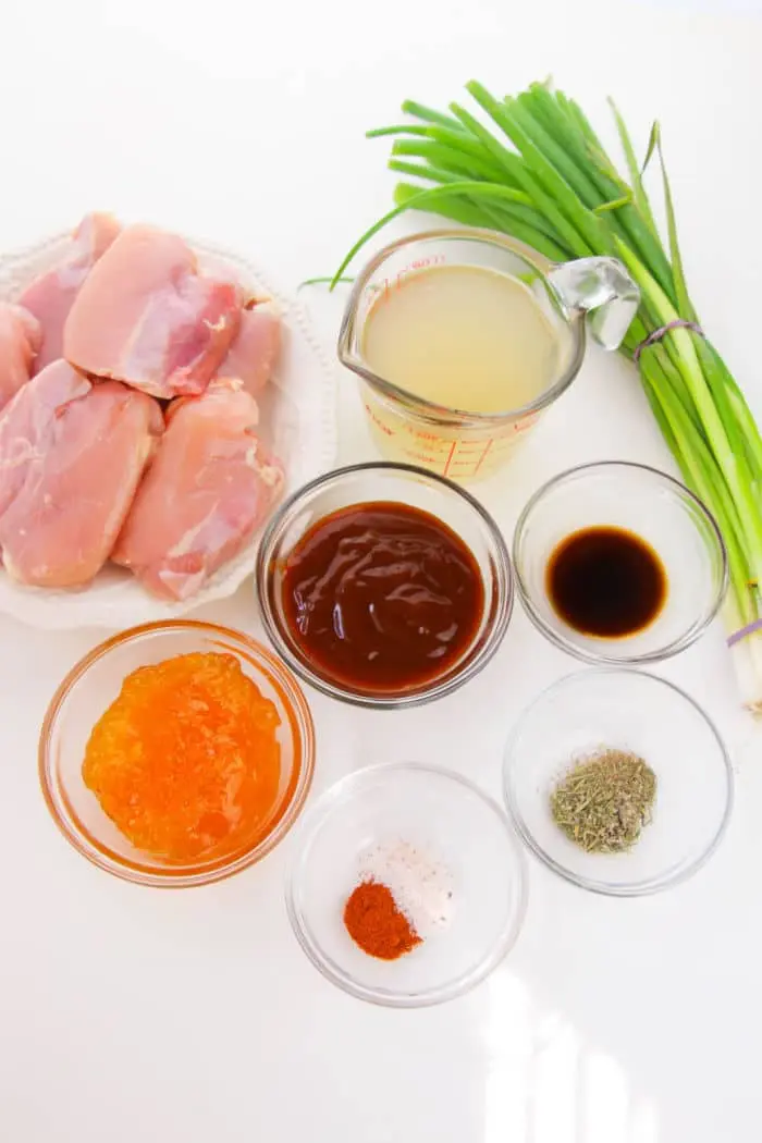 Ingredients on a white surface include boneless skinless chicken thighs on a plate, green onions tied with string, a measuring cup with liquid, small bowls of orange-colored sauce and brown sauce, soy sauce, and seasonings of salt, pepper, and paprika—perfect for your crockpot creation.