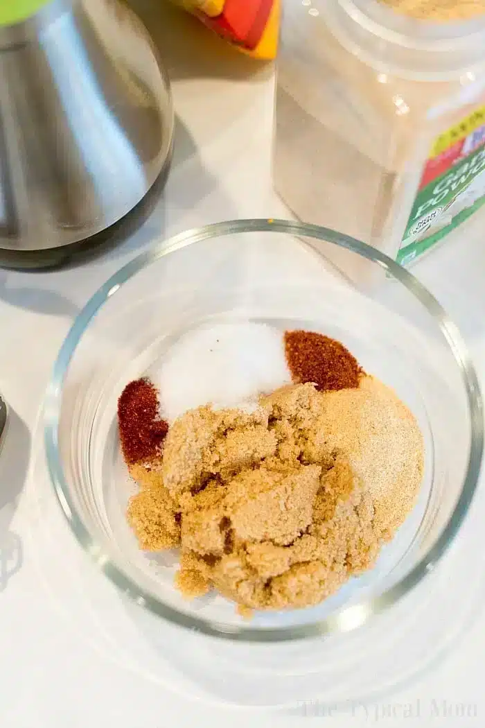 A glass bowl containing various seasonings, including brown sugar, garlic powder, paprika, and salt. A metal object and a container of spices are partially visible in the background on the white surface—perfect for flavoring bone-in air fryer chicken thighs.