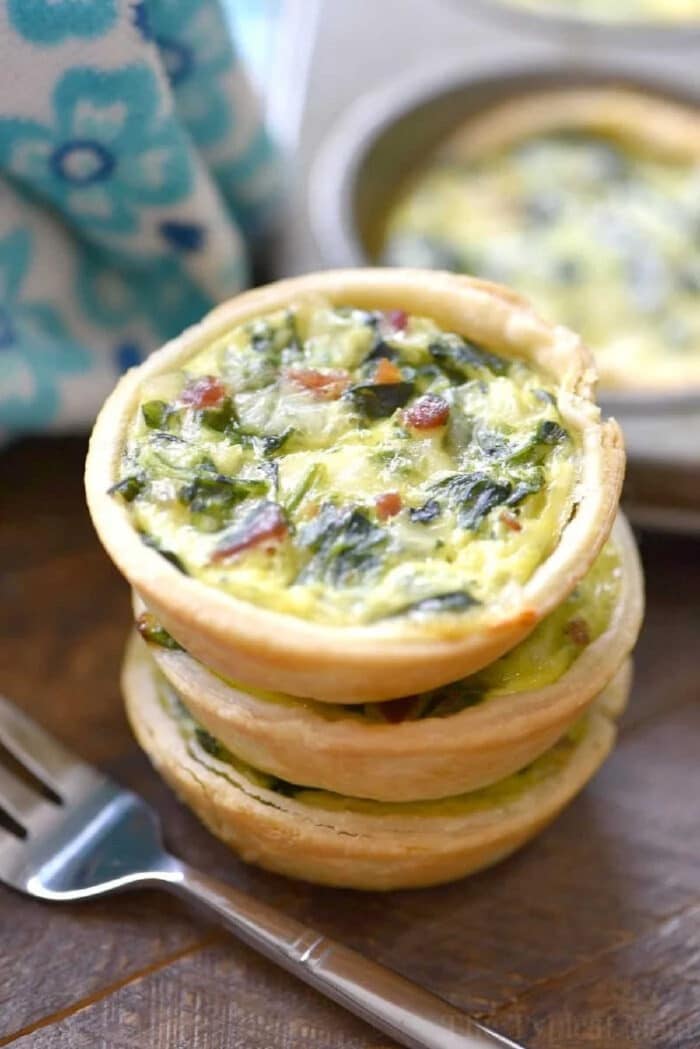 Three mini quiches made with an easy mini quiche recipe, featuring spinach and bacon, are stacked on a wooden table next to a fork and a blue patterned cloth.