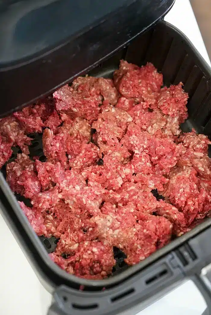 Ground beef is spread out in an air fryer basket, ready to be cooked. The air fryer lid is partially visible at the top of the image, perfect for those exploring new air fryer beef recipes.