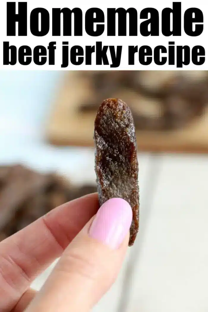 A close-up of a hand with pink-painted nails holding a piece of beef jerky. "Homemade air fryer beef jerky recipe" is written in bold black letters at the top of the image. In the background, more pieces of jerky are spread out on a wooden surface.