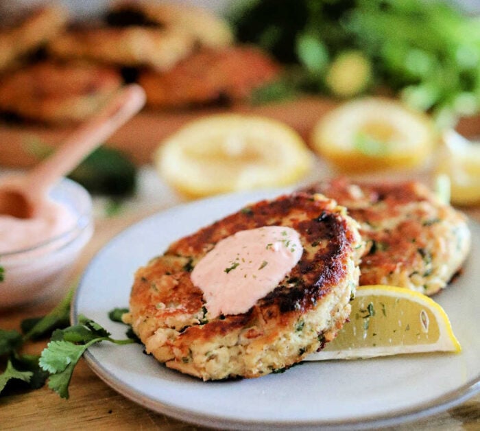 Two golden-brown patties with a dollop of sauce and a lemon wedge on a white plate, garnished with herbs—an appetizing presentation perfect for anyone curious about how to make tuna patties.