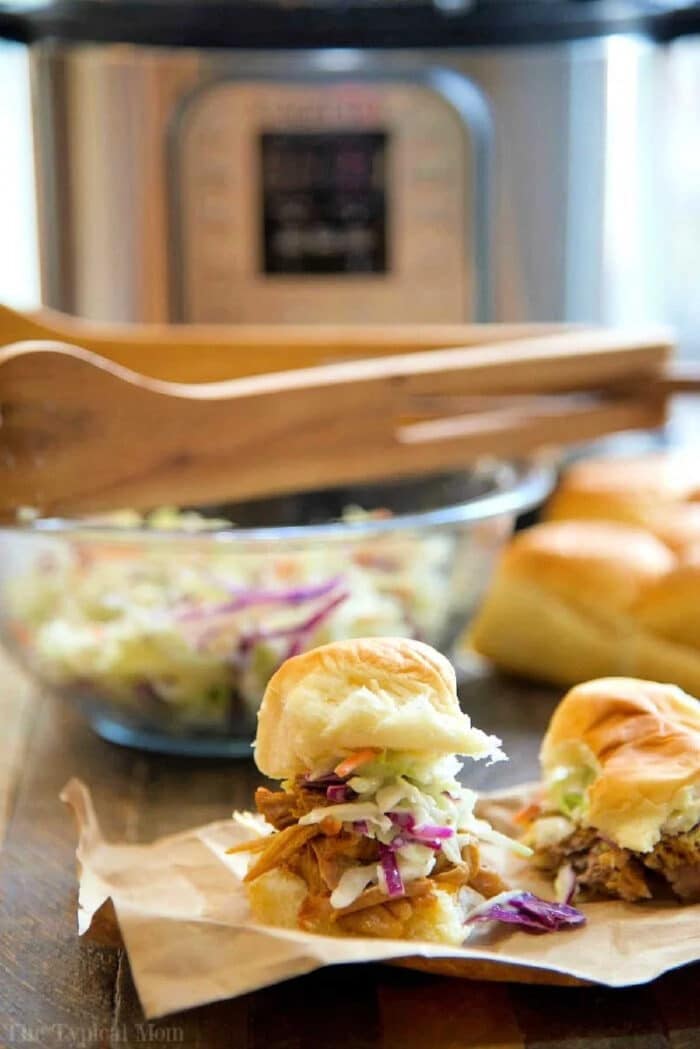 Easy Instant Pot pork shoulder recipe you will love. Amazing barbecue pulled pork sandwiches made in your pressure cooker that even my little one loves. Juicy pork usually takes all day to get this tender but in your Instant Pot it takes less than a few hours to fall apart. #pulledpork #pork #instantpot #pressurecooker #instantpotrecipes #thetypicalmom Pulled pork sliders topped with coleslaw on paper, with a bowl of slaw and a slow cooker in the background—perfect for serving after making an instant pot pork shoulder recipe.