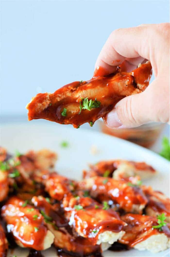 A hand holds a saucy chicken wing above a plate of barbecue wings, garnished with chopped herbs&mdash;perfect inspiration for making frozen chicken tenders in the Instant Pot at home.