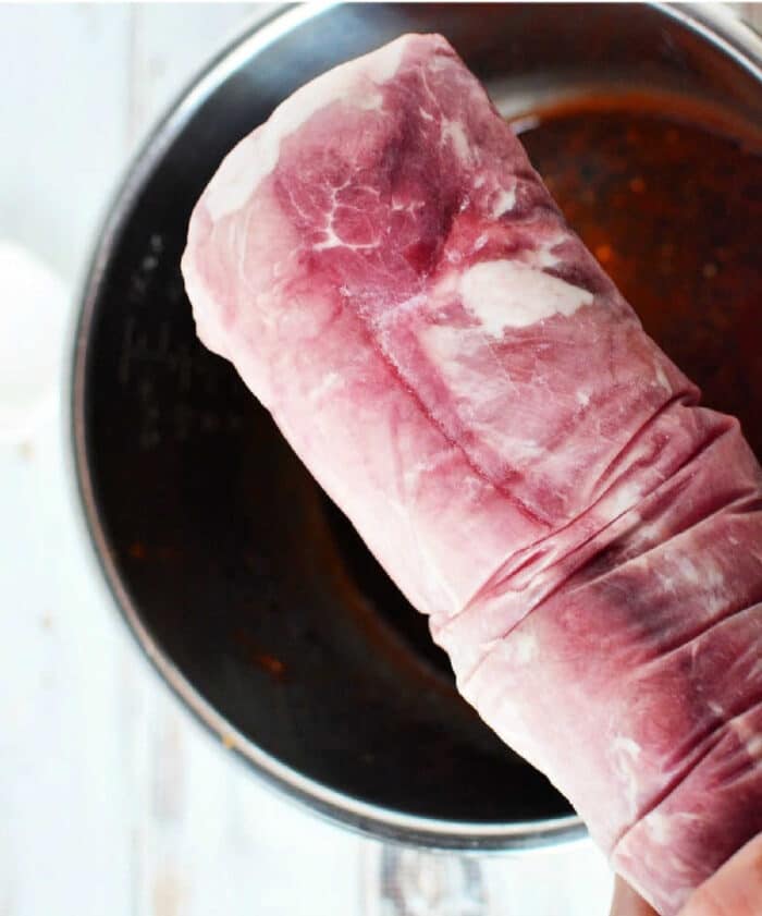 A raw pork tenderloin, perfect for making frozen pork loin in an Instant Pot, is held above a metal mixing bowl on a white surface.