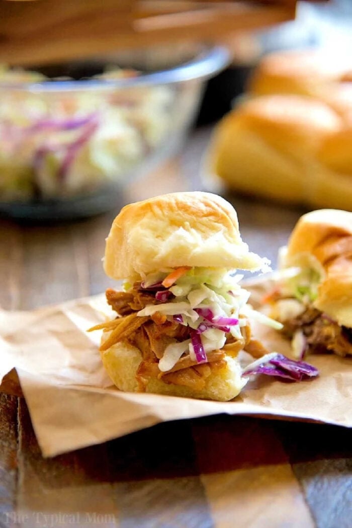 Instant Pot pork shoulder recipe you will love. Amazing barbecue pulled pork sandwiches made in your pressure cooker that even my little one loves. Juicy pork usually takes all day to get this tender but in your Instant Pot it takes less than a few hours to fall apart. #pulledpork #pork #instantpot #pressurecooker #instantpotrecipes #thetypicalmom Pulled pork slider topped with coleslaw on a bun, made using an instant pot pork shoulder recipe, placed on parchment paper with more sliders in the background.