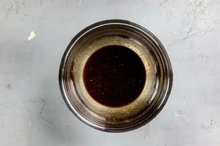 A stainless steel bowl containing a dark brown Asian Steak Marinade sits on a grey, textured surface.