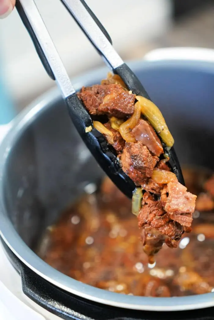 Tongs holding cooked pieces of beef and onions over a pot filled with broth suggest a delicious beef arm roast recipe. The meat appears tender, perfectly mixed with onions, indicating it’s been lovingly simmering in rich liquid.