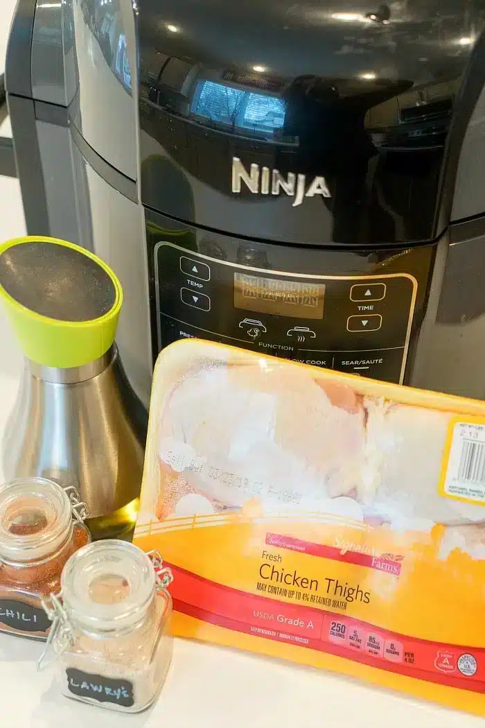 A Ninja countertop appliance is shown behind a package of uncooked fresh, bone-in, air fryer chicken thighs labeled Signature Farms. Two small spice jars labeled "Lawry's" and "Chili" and a stainless steel oil dispenser with a green top are also in the image.