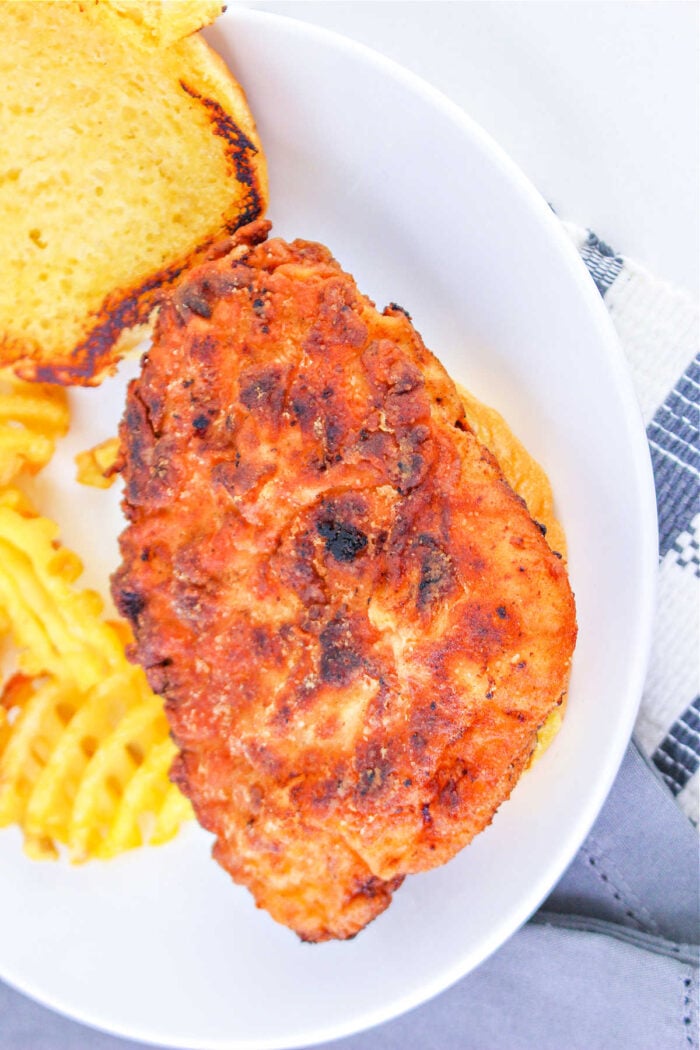 A fried chicken sandwich on a bun, perfectly crisp after learning how long to deep fry chicken breast, served with crinkle-cut fries on a white plate.