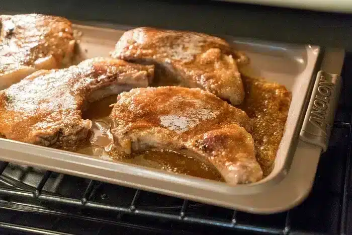 Baked pork chops on a metal baking tray inside an oven, showcasing the best baked pork chops. Coated with a golden brown seasoning and surrounded by their juices, the tray features a handle with the brand name "Anolon" visible.