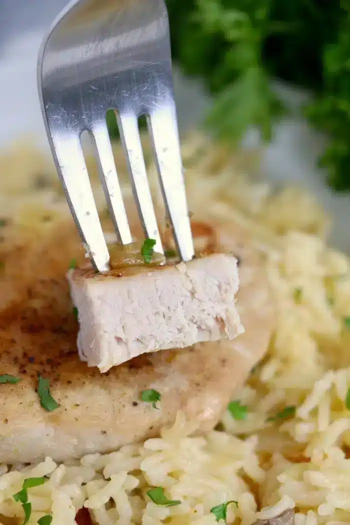 A fork holding a piece of cooked chicken over a plate of seasoned rice garnished with chopped herbs, evoking flavors as delightful as the best baked pork chops. A sprig of fresh parsley is visible in the background.