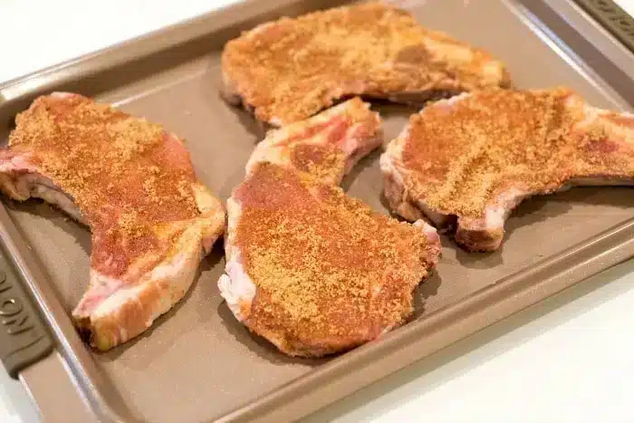 Four seasoned pork chops arranged on a baking sheet, ready for cooking. The chops, promising to be the best baked pork chops you've tasted, are sprinkled with a brown spice mix. The baking sheet is placed on a light-colored surface.